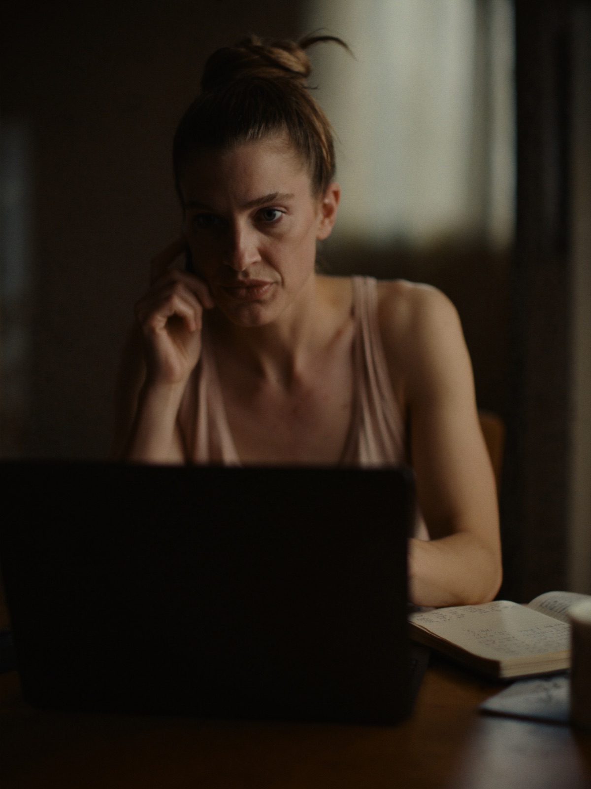 A woman on the phone at her table in the evening, laptop open, listening closely