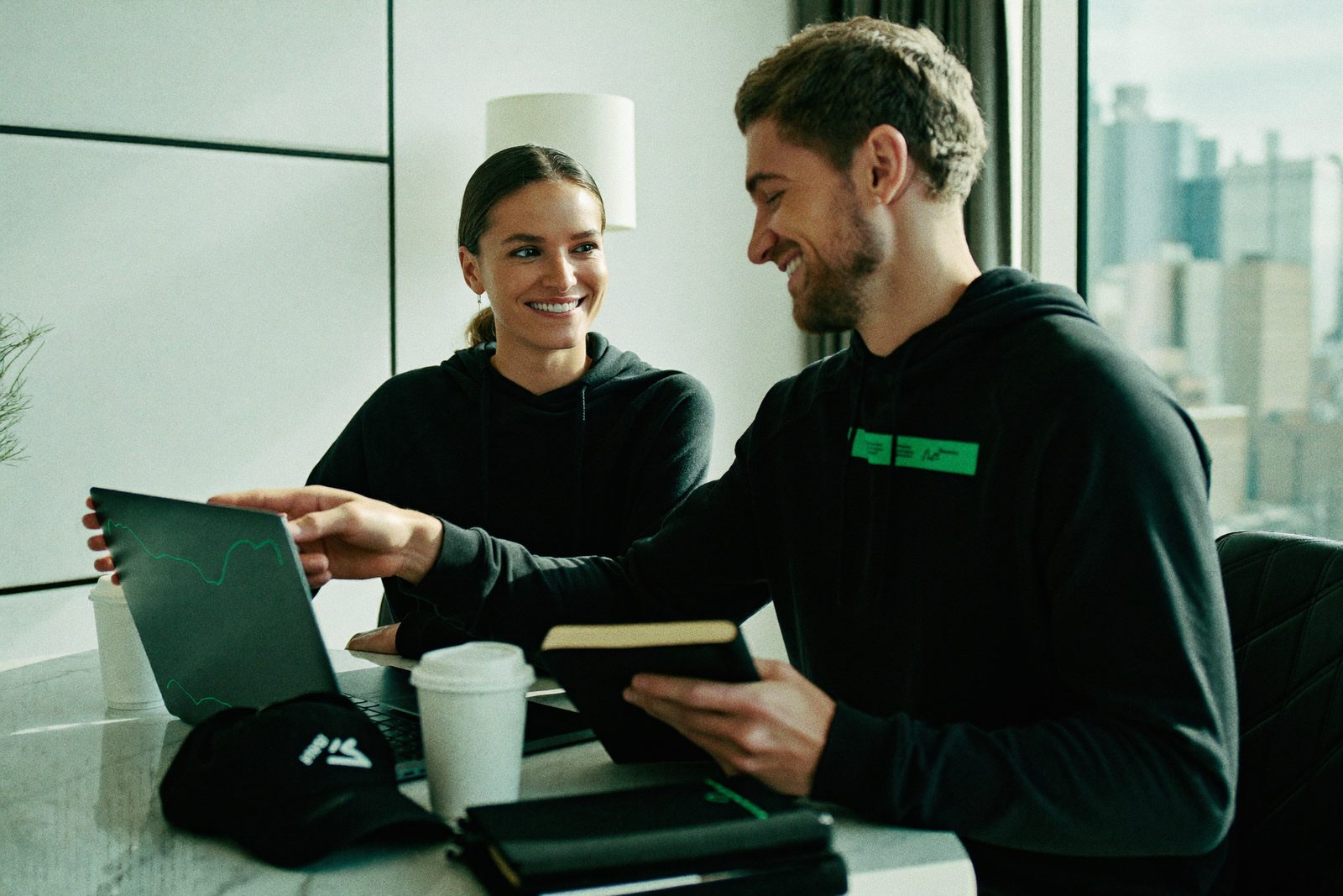 Two members at a laptop in a bright hotel room, one explaining a chart to the other.